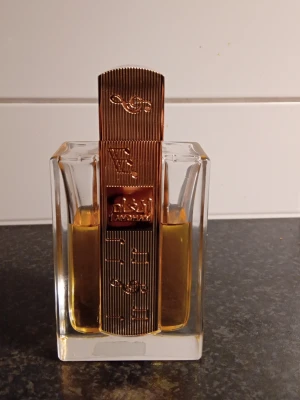 Lattafa Angham Eau de Parfum 100ml - Exklusiv parfym från Lattafa, Angham, i en elegant glasflaska med guldfärgad dekor och lock. Flaskan har en modern, fyrkantig form och doften kommer i en lyxig beige förpackning med gulddetaljer. Innehåller 100 ml Eau de Parfum. Den kommer helt ny kommer med förpackningen 