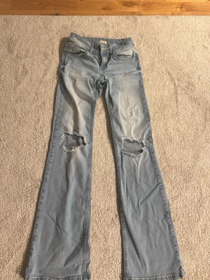 Ljusblå bootcut jeans med hål från Nelly - Säljer ett par ljusblå bootcut jeans med slitna hål på båda knäna. Jeansen har klassisk femficksdesign och är tillverkade i bomull. Perfekta för en avslappnad och trendig look. Storlek S! 