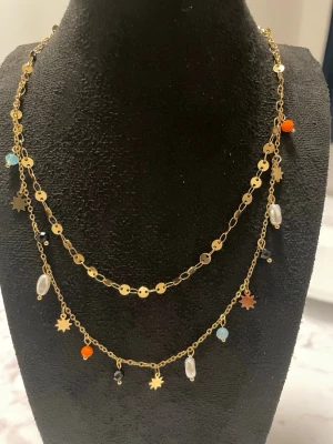Guldigt & rostfritt  halsband med pärlor och stjärnor - Dubbelradigt halsband i guldfärgad metall med små stjärnhängen och färgglada pärlor i orange, blått, svart och vitt. Kedjan har dekorativa runda länkar och detaljer som ger en trendig och lekfull look. Materialet är i Copper och rostfritt. Flera hackar för längd justering. Längd 34cm+5 cm hackarna 