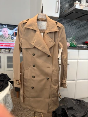 Beige trenchcoat från Charles Jourdan - Stilren beige trenchcoat från Charles Jourdan med klassisk dubbelknäppning, bred krage och bälte i midjan. Jackan har axelklaffar, justerbara ärmslut och två sidofickor. Perfekt för dig som vill ha en tidlös och elegant look.