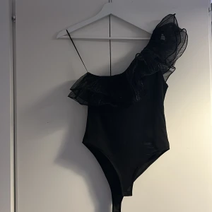 Svart oneshoulder body från Zara - Säljer en svart body från Zara med en axelbandslös design och volang i transparent tyg över ena axeln. Ena axeln har smalt band och den andra är oneshoulder med stor volangdetalj. Perfekt för dig som vill sticka ut med en cool och elegant look. Stringbody. 