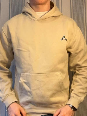 Beige hoodie från Jordan med logga - Snygg beige hoodie från Jordan med klassisk svart logga på bröstet. Hoodien har känguruficka framtill och en skön huva. Perfekt för en chill och avslappnad stil. Tillverkad i mjuk bomullsmix för extra komfort.
