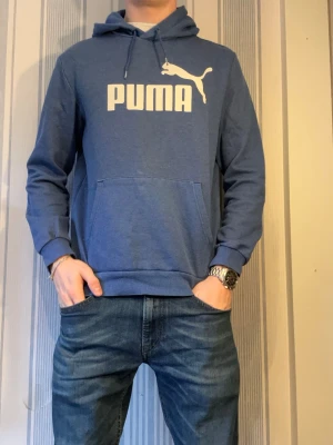 Blå hoodie från Puma med logga - Snygg blå hoodie från Puma med stor vit logga på bröstet. Klassisk modell med huva och dragsnören samt en stor magficka framtill. Perfekt för en avslappnad och sportig stil. Mjuk bomullsblandning och ribbade muddar vid ärmslut och nederkant.