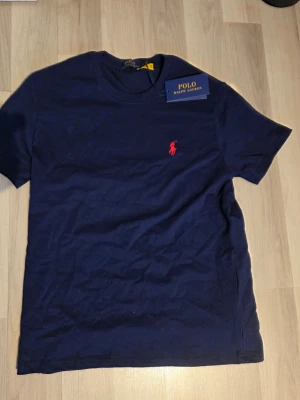 Marinblå t-shirt från Polo Ralph Lauren - Snygg marinblå t-shirt från Polo Ralph Lauren med klassisk röd logga broderad på bröstet. Tillverkad i mjuk bomull och har en normal passform med korta ärmar och rund hals. Perfekt basplagg för en stilren look.