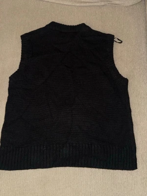 Svart stickad väst med ribbade kanter - En svart stickad väst med ribbade kanter vid hals, ärmhål och nederkant. Klassisk och enkel design som passar perfekt över en skjorta eller t-shirt. Västen har en rak passform och är superenkel att styla för en trendig look.