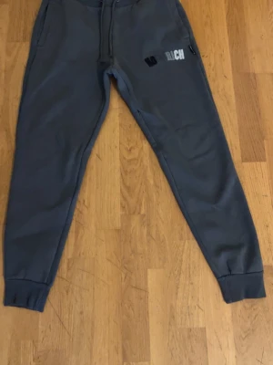 Gråa mjukisbyxor från Hoodrich - Mörkgrå mjukisbyxor från Hoodrich med broderad logga på ena benet. Byxorna har snörning i midjan, ribbade muddar vid bensluten och praktiska sidofickor. Perfekta för en avslappnad och stilren look. Skicket är använd och där med priset lågt. 