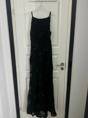 Gina långklänning  - Elegant svart långklänning med tunna axelband och diskret blommönster i tyget. Klänningen har en figurnära passform upptill och faller snyggt längs kroppen. Perfekt för dig som vill ha en stilren och klassisk look.