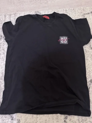 Svart t-shirt från Hugo med tryck - Säljer en svart t-shirt från Hugo med ett litet tryck på bröstet där det står 'HUGO' i vitt och rött. Klassisk passform och rund halsringning. Perfekt till jeans eller shorts för en clean och enkel stil.