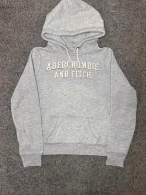 Sällsynt Grå vintage hoodie från Abercrombie & Fitch - Ljusgrå hoodie med vit broderad text 'Abercrombie and Fitch' på bröstet. Klassisk känguruficka framtill, dragsko i huvan och mjukt material. Perfekt för en avslappnad och stilren look.