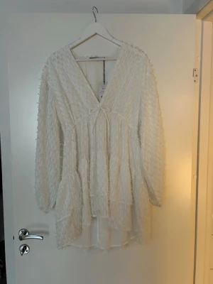 Vit omlottklänning med volang - Superfin vit klänning med v-ringning, omlottkänsla och volangdetaljer. Klänningen har lång ärm och en liten rosett framtill. Perfekt till studenten eller en somrig sommarklänning 