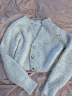 Ljusgrå fluffig kofta med pärlknappar - Supermjuk och fluffig ljusgrå kofta med v-ringning och tre snygga pärlknappar med gulddetaljer. Kortare modell med långa ärmar och ribbade muddar. Perfekt för lager-på-lager och en mysig look. Storlek M men skulle säg att det passar någon med en Xs/s