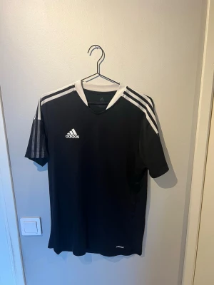 Svart Adidas sporttröja  - Svart sporttröja från Adidas i storlek M med vita detaljer och klassiska tre ränder på axlarna. Tröjan är tillverkad i ett lätt och snabbtorkande material, perfekt för träning eller fotboll.
