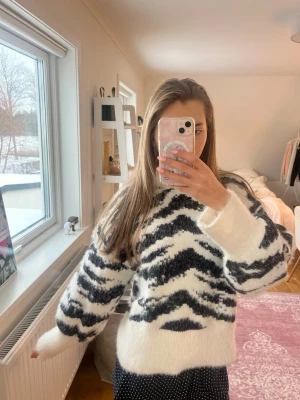 Zebramönstrad fluffig stickad tröja - Säljer en vit och svart zebramönstrad stickad tröja med oversized passform och långa ärmar. Tröjan är fluffig och mjuk, perfekt för att hålla sig varm. Materialet ser ut att vara en blandning med mohair eller liknande. Snygg och trendig design.