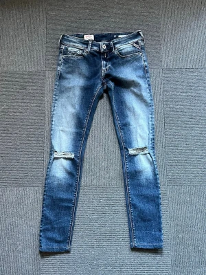 Blå slitna skinny jeans med hål - Säljer ett par blå skinny jeans från replay  med slitningar och hål vid knäna. Jeansen har klassisk femficksmodell, dragkedja och knapp i midjan samt ljusare tvättade partier för en trendig look. Perfekta för dig som gillar en avslappnad och cool stil. Storlek 25/30
