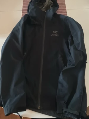 Svart regnjacka från Arc'teryx - Snygg svart regnjacka från Arc'teryx med huva och vattentäta dragkedjor. Defekter är den har lite slit på vänster höft sida sen vita små prickar på bröstet