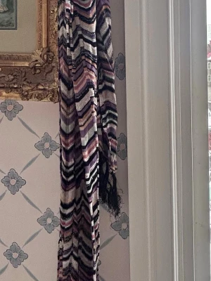 Mönstrad halsduk i pastell och svart - Säljer en lång, stickad halsduk med zigzag-mönster i lila, svart, beige, vitt, guld och rosa. Halsduken har tunna glittriga inslag och fransar i ändarna. Perfekt för dig som gillar färg och retrovibbar. Missoni by Lindex 