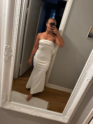 Vit axelbandslös långklänning - Säljer en elegant vit långklänning med axelbandslös design och draperad detalj över bröstet. Klänningen har en slank passform och faller snyggt längs kroppen. Perfekt för dig som vill ha en stilren och modern look.