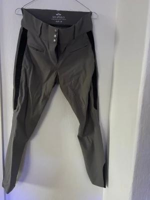 Grå ridbyxor från HV Polo - Snygga grå ridbyxor från HV Polo med hög midja, fyra silverknappar och förstärkta partier på insidan av benen. Byxorna har två fickor fram och svarta detaljer längs sidorna och vid bensluten. Perfekta för ridning och träning. Lite sömsläpp i överkant 