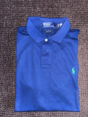 Ralph Lauren Polo - Snygg blå pikétröja från Ralph Lauren med klassisk krage och två knappar. Tröjan har den ikoniska gröna logotypen broderad på bröstet och är tillverkad i bomull. Perfekt för en clean och stilren look. Hem utan rök och djur. Endast använd ett par gånger och i nyskick. Skickas omedelbart. Just nu fri frakt!