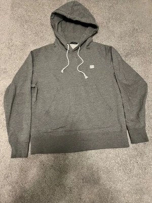 Grå hoodie från Acne Studios - Säljer en stilren grå hoodie från Acne Studios med vit dragsko i huvan och den klassiska lilla Acne-loggan på bröstet. Hoodien har en avslappnad passform och är tillverkad i mjuk bomullsblandning. Perfekt för en casual och trendig look.