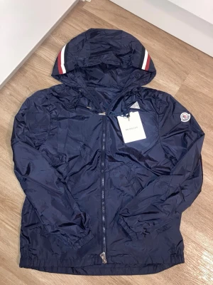 Mörkblå vindjacka från Moncl3r - Snygg mörkblå vindjacka med huva och dragkedja framtill. Jackan har vita och röda ränder på huvan samt logga på ärmen. Tillverkad i lätt, glansigt material och har elastiska muddar vid ärmsluten.