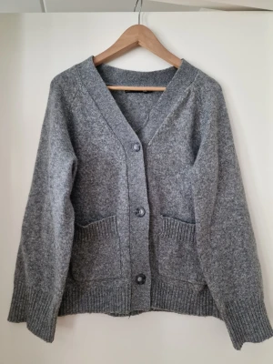 Grå stickad kofta med fickor - Mysig grå stickad kofta med v-ringning och stora knappar framtill. Koftan har två rymliga fickor och ribbade muddar vid ärmslut och nederkant. Perfekt för lager på lager och enkel att matcha med olika outfits. Storlek L. Fint skick