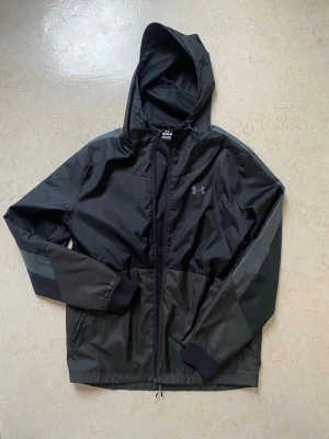 Svart vindjacka från Under Armour - Snygg svart vindjacka från Under Armour med huva och dragkedja framtill. Jackan har diskreta grå detaljer på ärmarna och elastiska muddar vid handlederna. Perfekt för blåsiga dagar och sportiga outfits. Liten logga på bröstet och lätt material.