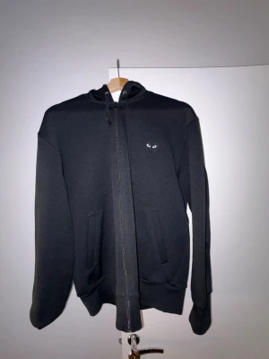 Svart hoodie Comme des Garçons Play - Svart hoodie med dragkedja från Comme des Garçons Play. Har huva med dragsko, två fickor fram och det ikoniska hjärtat med ögon på ryggen samt ett litet hjärta på bröstet. Perfekt för en stilren och cool look.