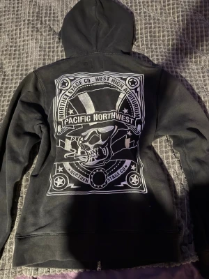 Svart hoodie vailent dreaming in scandinavia - Svart hoodie med dragkedja från vailent dreaming in scandinavia. Stor tryckt design på ryggen med texten 'Pacific Northwest' och en döskalle med hatt. Två fickor fram och huva. Perfekt för dig som gillar streetwear och coola tryck.