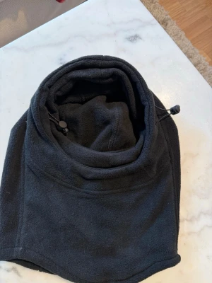 Svart fleecehuva med dragsko - Säljer en svart huva i mjuk fleece med justerbar dragsko. Perfekt att dra över huvudet för extra värme och skydd mot vind. Enkel att använda och passar till de flesta jackor och tröjor. Stilren design utan synligt märke.