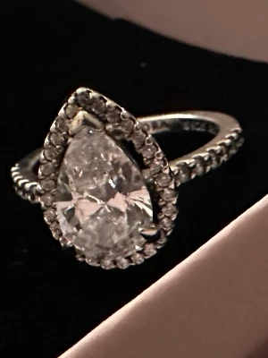 PANDORA - Snygg silverfärgad ring med en stor, droppformad klar sten i mitten, omgiven av mindre gnistrande stenar runt hela stenen och längs bandet. Perfekt för dig som gillar statement-smycken och vill ha något som verkligen syns.