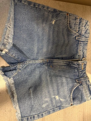 Blå jeansshorts med fransig kant - Säljer ett par klassiska blå jeansshorts med fransig kant och slitna detaljer. Shortsen har fem fickor, dragkedja och knapp framtill. Perfekta för sommaren och ger en avslappnad vibe.