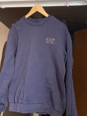 Blå tracksuit från EA7 Emporio Armani - Mörkblå sweatshirt från EA7 Emporio Armani med rund hals och tryckt logga på bröstet. Det är storlek L men passar dig som är ca 180 cm väldigt bra.  Perfekt för en sportig och chill look. Pris kan diskuteras!