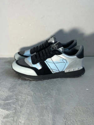 Valentino Garavani blå sneakers - Snygga sneakers från Valentino Garavani med blå, grå och svarta kamouflagemönster. Skorna har svarta snören, detaljer i mocka och läder samt nitar på hälen. Insidan är vadderad för extra komfort och sulan är svart.