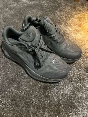 Mörkgrå sneakers från Alo - Snygga mörkgrå sneakers från Alo med modern design och diskreta loggor på plös och tå. Ovandelen är i mesh för bra andningsförmåga och sulan är robust och bekväm. Perfekta för en sportig och stilren look.