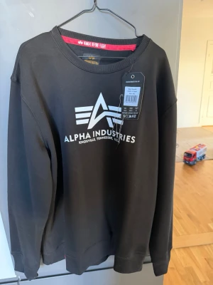 Svart Alpha Industries sweatshirt - Svart sweatshirt från Alpha Industries med vit logga och text på bröstet. Tröjan har rund halsringning, ribbade muddar och är tillverkad i mjukt material. Perfekt för en avslappnad och cool stil. Provad men aldrig använd. Något liten i storlek.