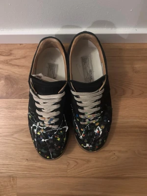 Svarta sneakers från Maison Margiela - Unika svarta sneakers från Maison Margiela med färgstänk i vitt, gult, blått och rött över hela ovandelen. Skorna har beige snörning och en klassisk låg siluett. Insidan är ljus och materialet ser ut att vara en mix av mocka och läder. Perfekt för dig som gillar statement-plagg.
