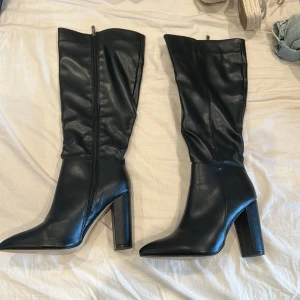 Svarta höga stövlar med klack - Snygga svarta stövlar i skinnimitation med spetsig tå och hög blockklack. Stövlarna har dragkedja på insidan och en stilren, slät design som ger en edgy vibe. Perfekta till jeans eller kjol för en trendig look.