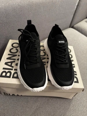 Helt Ny Bianco Sneakers, Svart, EUR 36. (37 /37,5)   - Snygga svarta sneakers från Bianco med vit sula och mesh-överdel. Skorna har en sportig look med klassisk snörning och låg profil. Perfekta för dig som gillar stilrena och bekväma sneakers till vardags. Materialet är en kombination av textil och syntet.