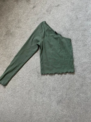 Grön oneshoulder topp - Trendig grön oneshoulder topp med lång ärm och croppad passform. Toppen har vågig kant nertill och i ärmarna, säljer då den är för liten. Är helt ny och oanvänd! 