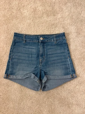 Blå denimshorts med uppvik - Klassiska blå jeansshorts med uppvikta benslut och femficksdesign. De har normal passform, bälteshällor och stängs med knapp och dragkedja. 
