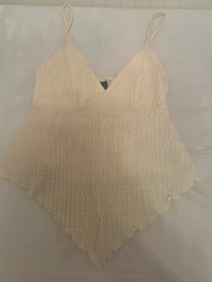 Beige linnetopp med smala axelband - Supersöt beige linnetopp med smala axelband och v-ringning. Toppen har en lätt skrynklig struktur och spetsig nederkant både fram och bak, vilket ger en unik och trendig look. Perfekt för varma dagar och enkel att styla till olika outfits.