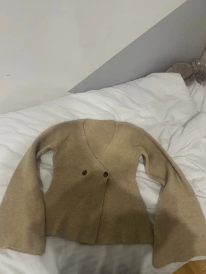 Beige ribbstickad omlottkofta - Snygg beige ribbstickad omlottkofta med två knappar framtill och vida ärmar. Koftan har en djup V-ringning och är gjord i ett mjukt stickat material som känns riktigt skönt mot huden. Perfekt för lager-på-lager och chill dagar.