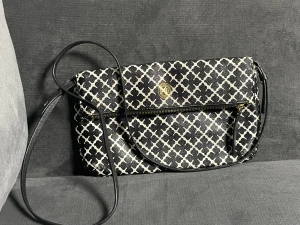 Svartvit axelväska med mönster - Snygg svartvit axelväska med grafiskt rutigt mönster och guldfärgad detalj framtill. Väskan har dragkedja och lång axelrem. Tillverkad i syntetmaterial och har ljus insida. Perfekt för att bära det viktigaste med stil. Obs, lite kedje lite lite trasig