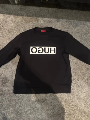 Svart sweatshirt från Hugo Boss - Cool svart sweatshirt från Hugo Boss med vit spegelvänd HUGO-logga på bröstet. Tröjan har rund halsringning och långa ärmar. Perfekt för en stilren och trendig look.