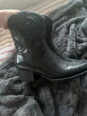 Svarta cowboy boots från Duffy - Snygga svarta cowboy boots från Duffy med klassisk spetsig tå och bred klack. Skorna har dekorativa sömmar och är tillverkade i ett slätt, konstläderliknande material. Dragkedja på insidan för enkel påtagning. Perfekta för dig som vill ha westernstil.