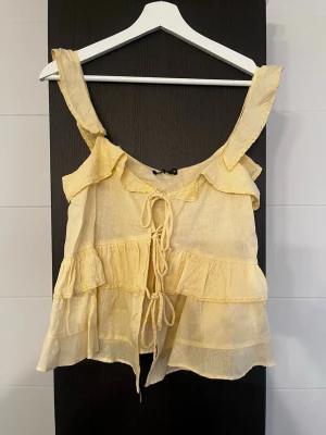 Zara söt topp i storlek XS - Supersöt ljusgul linnetopp från ZARA med volangdetaljer och knyt framtill. Toppen är ärmlös med breda axelband och har en luftig, flowy passform. Perfekt för varma dagar och ger en romantisk vibe till din outfit. Ny med prislapp