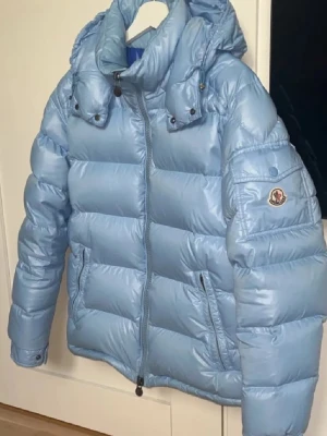 Ljusblå Moncler jacka - Säljer en ljusblå pufferjacka från Moncler med huva och dragkedja framtill. Jackan har ett glansigt yttermaterial och ikonisk Moncler-logga på ärmen. Perfekt för kalla dagar och har en riktigt snygg streetstil. Flera fickor med dragkedja och quiltad design. Säljer då den ej passar och köpte fel storlek 