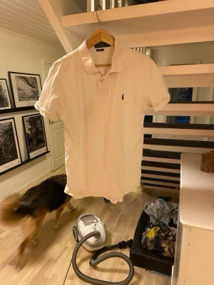 Vit piké från Polo Ralph Lauren - Klassisk vit pikétröja från Polo Ralph Lauren med liten broderad logga på bröstet. Tröjan har krage, knappar vid halsen och korta ärmar. Tillverkad i mjuk bomull för en bekväm känsla och clean look. Liten fläck men syns knappt.