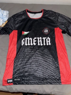 Svart och röd Omerta matchtröja - Säljer en svart och röd matchtröja från Omerta med vita detaljer och tryck. Tröjan har nummer 47 på ryggen, logga på bröstet och coolt mönster i tyget. Tillverkad i lätt och ventilerande material, perfekt för träning eller match.